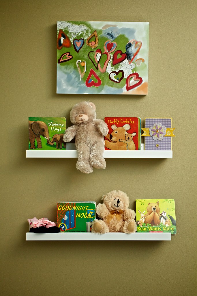 Wall-mounted nursery bookshelves with children's books, stuffed teddy bears, and a pair of pink baby shoes, with a colorful heart-themed painting above.