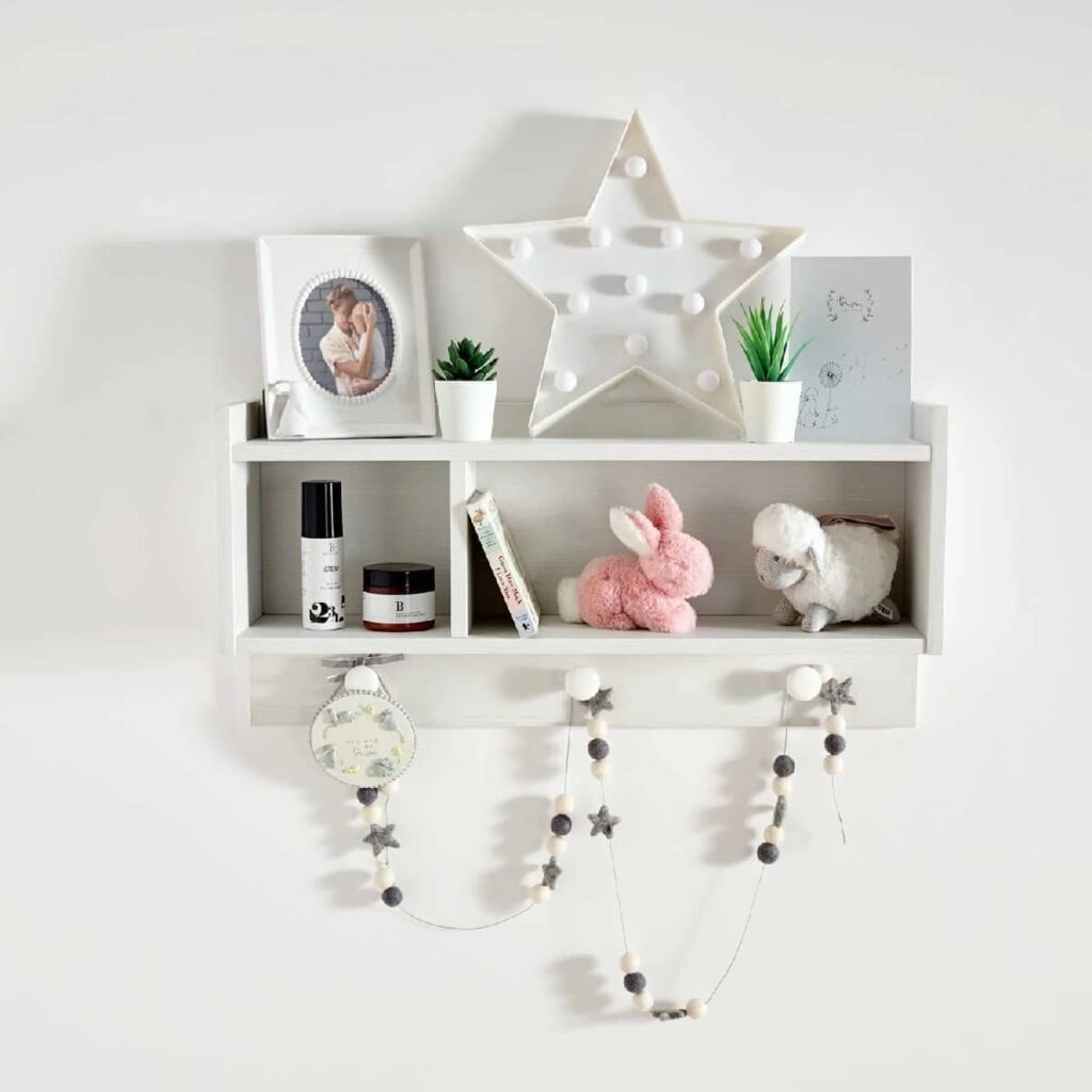 White wall-mounted nursery book shelves with decorative items including a framed photo, star-shaped light, small plants, a pink stuffed bunny, a white stuffed lamb, and string lights with stars hanging below.