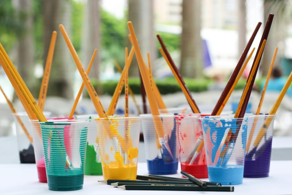 Plastic cups filled with various colored paints and paintbrushes on a table, with a background of trees and a blurred outdoor setting.