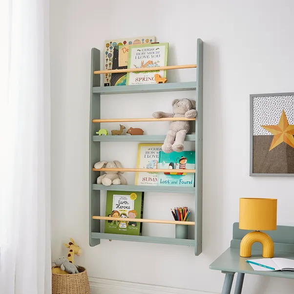 A child's room with a light blue and yellow wooden floating nursery book shelves mounted on the wall, holding books, a stuffed animal, and small toys. A desk with a lamp and colorful pencils is next to the bookshelf, and a star-themed wall art piece is visible.