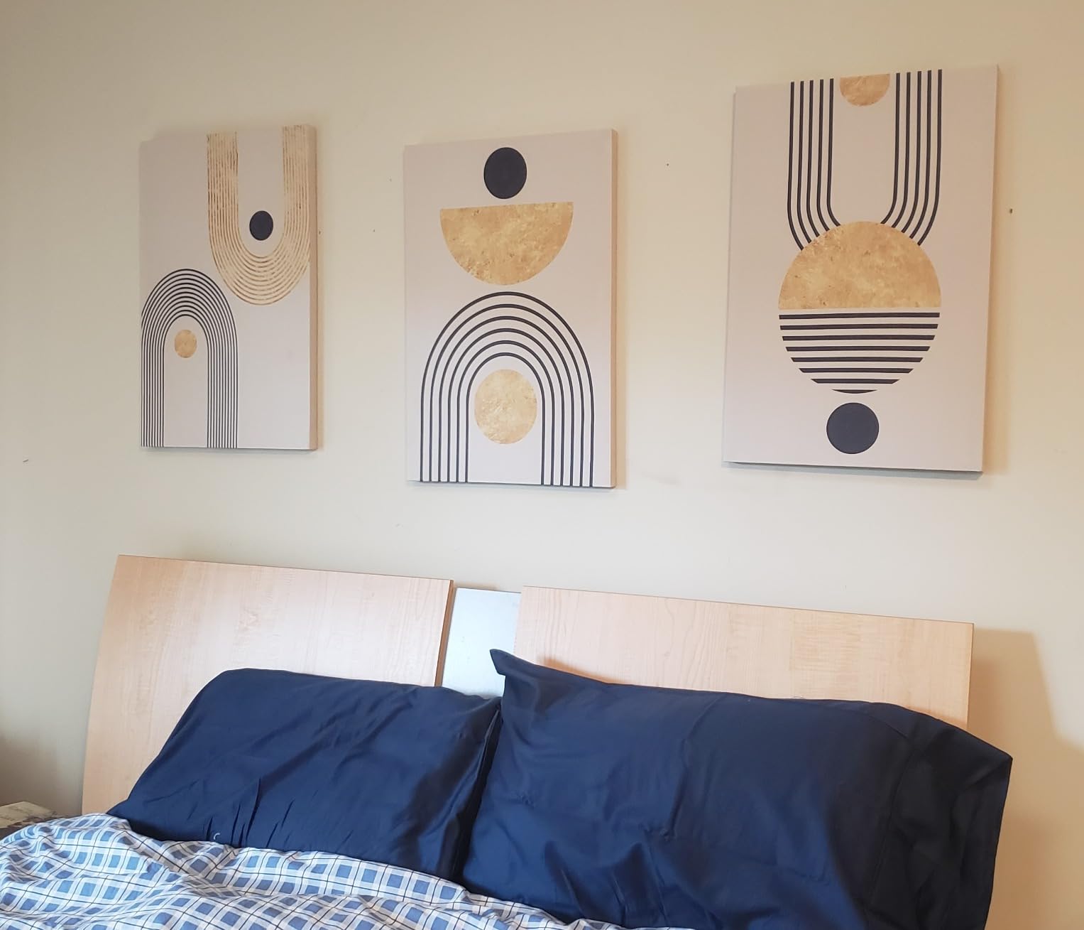 Bedroom with a wooden headboard and blue pillows, featuring three abstract art canvases with geometric designs in gold and navy blue, including circles, semi-circles, and arches.