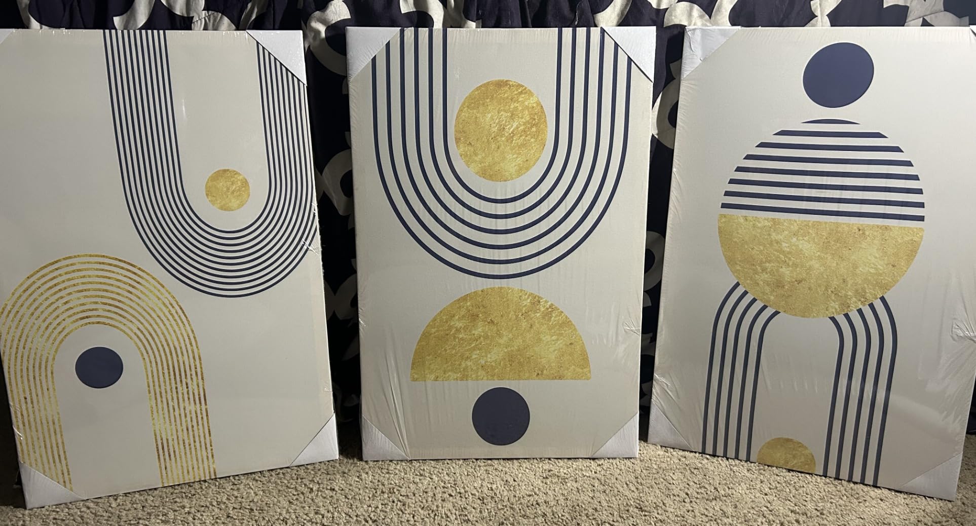 Three abstract art canvases with geometric designs, featuring gold and navy blue shapes and lines. The artwork includes circles, semi-circles, and arches, still wrapped in plastic.