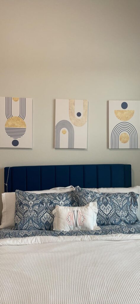 Bedroom with a blue and white color scheme, featuring three abstract art canvases above the bed. The artwork has geometric designs in gold and navy blue, with circles and arches.