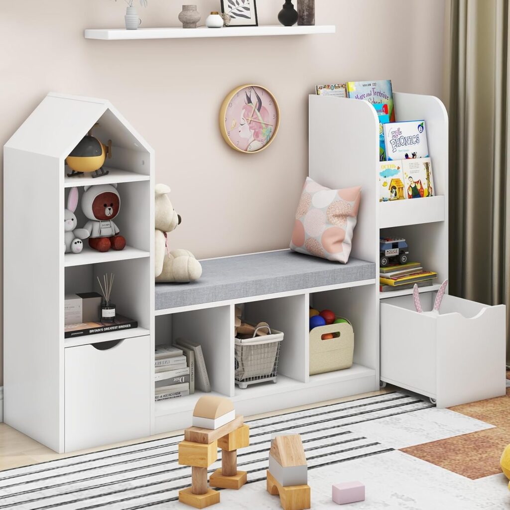 A children's room with a white and gray storage bench, featuring open shelves filled with toys, books, and a pink pillow. Above the bench, a white wall shelf holds small decorative items, and a wall clock is mounted nearby.