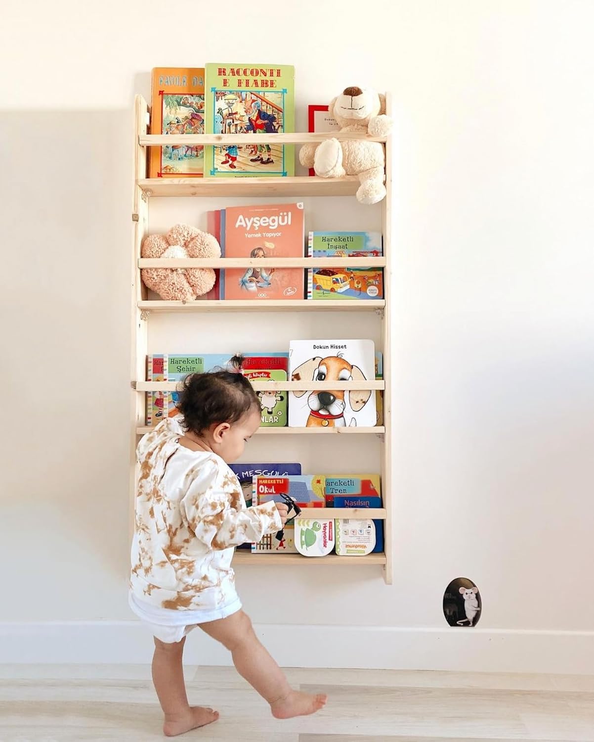 A toddler standing in front of a wall-mounted wooden bookshelf filled with colorful children's books and soft toys. The child is reaching for a book on the lower shelf.