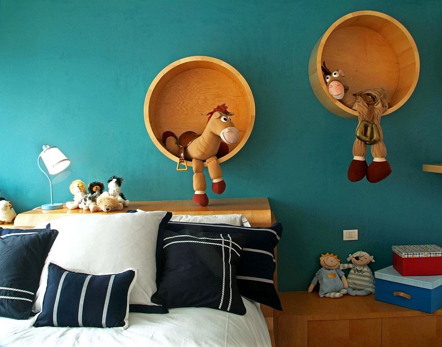 A children's bedroom with teal walls featuring two circular wooden shelves holding plush toy horses. The bed below has white and navy blue bedding, with several stuffed animals placed on it.