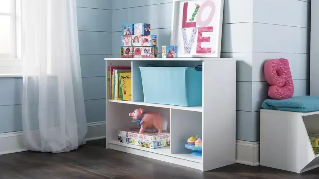 A white shelving unit in a child's room, containing books, toys, and storage bins. A framed print with the word "LOVE" is placed on top of the shelf, and a window with white curtains is visible in the background.