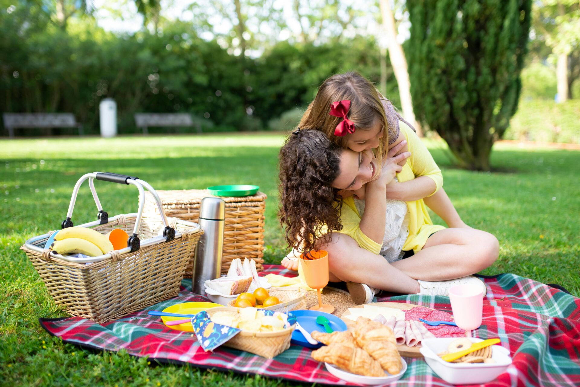 A mother and daughter enjoying a picnic on a grassy field. The picnic spread includes a wicker basket, sandwiches, croissants, fruit, and other snacks on a red checkered blanket. The daughter hugs the mother from behind, both smiling and dressed in yellow.