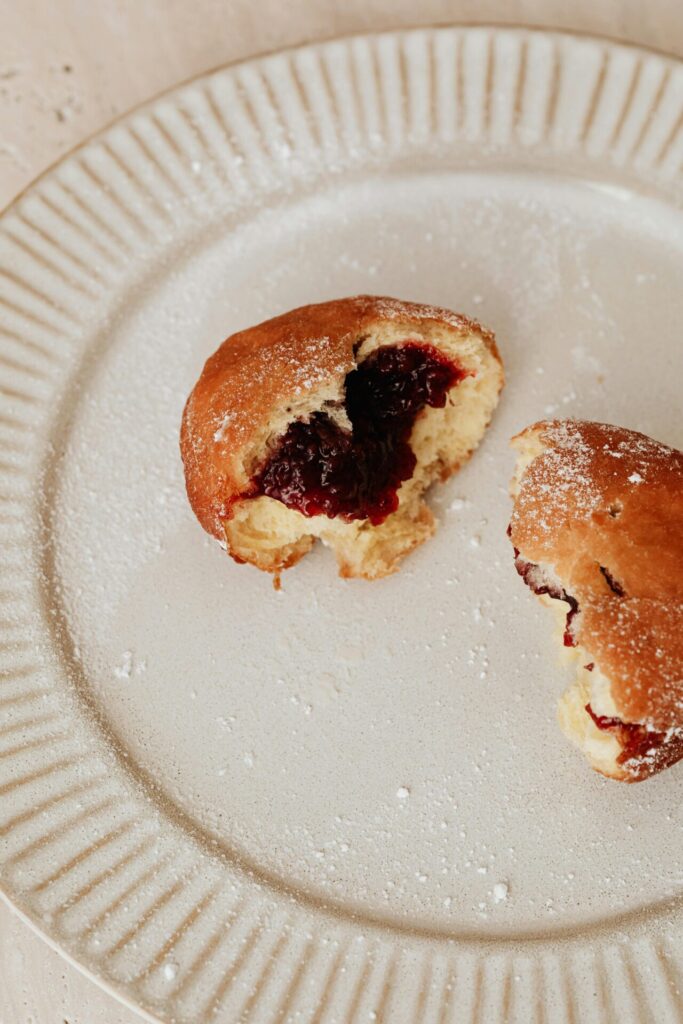 A jelly-filled donut, cut in half, placed on a beige ceramic plate dusted with powdered sugar.