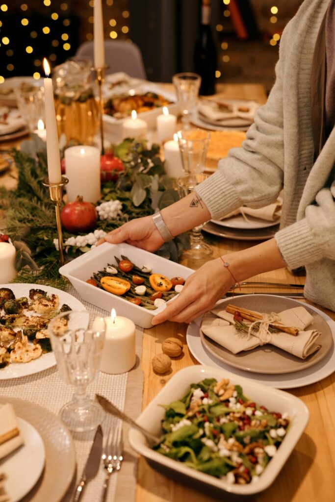 A festive dinner table setting with various dishes, candles, and decorations. A person in a gray sweater is placing a white serving dish filled with roasted vegetables on the table.