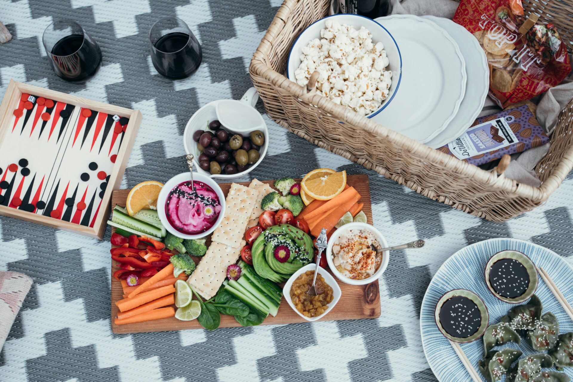A picnic setup on a patterned blanket featuring a colorful assortment of snacks including sliced vegetables, dips, olives, popcorn, and crackers. There is also a backgammon board and two glasses of red wine.