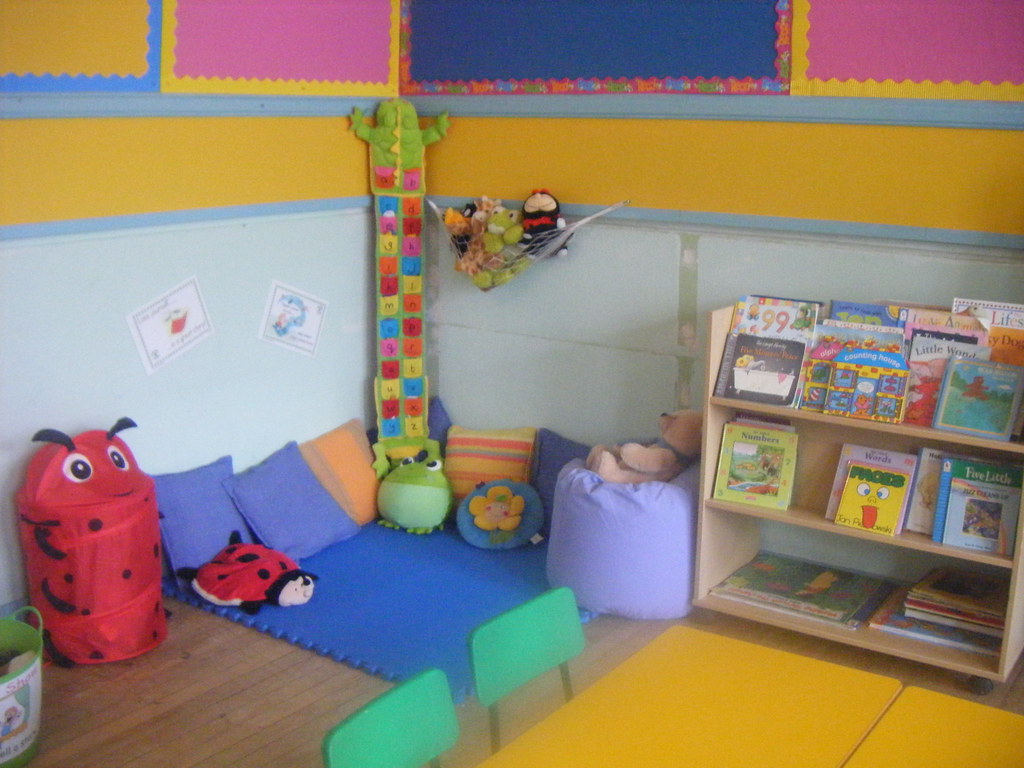 A colorful reading nook in a nursery, featuring a padded play area with pillows, stuffed animals, and a tall, playful nursery book shelf filled with children's books.