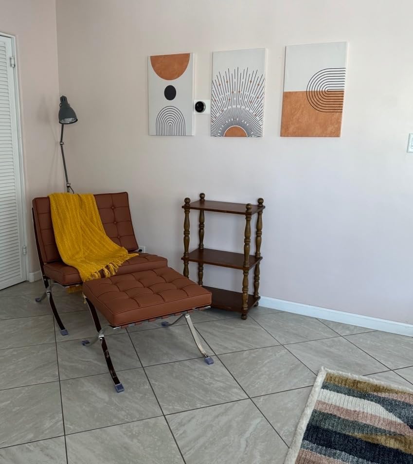 A bright room featuring a brown leather chair with a yellow throw blanket, a small wooden shelf unit, and three pieces of bohemian wall decor with abstract designs in orange and black hanging on the wall.