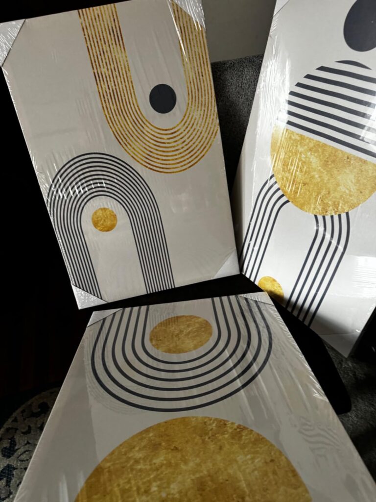 Three pieces of bohemian wall decor with abstract geometric designs in navy and gold, still wrapped in plastic, leaning against a dark surface.