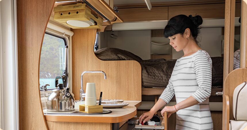 A woman organizing a drawer in a compact kitchen inside an RV, with a paper plate dispenser mounted underneath the overhead cabinet on the counter.