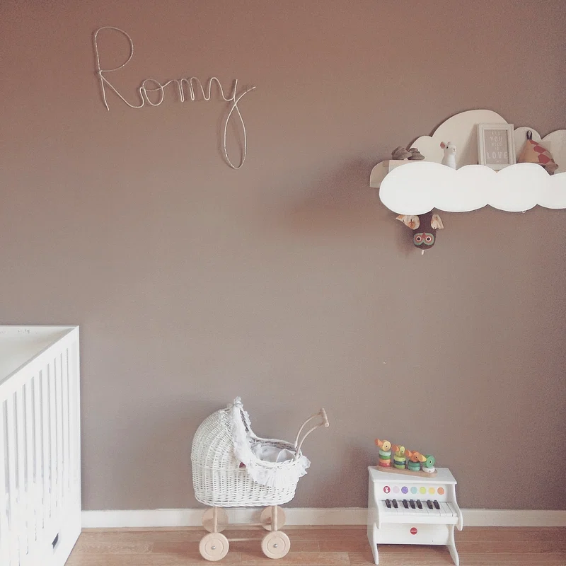 A nursery room with the name Romy artistically written on the wall in wire. A nursery book shelf in the shape of a cloud holds toys, while a white wicker baby carriage and a small toy piano sit below.