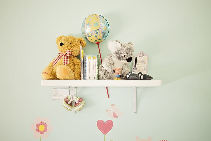 A nursery book shelf mounted on a pastel-colored wall, holding stuffed animals, baby books, and a balloon with 'It's a Boy!' text, creating a cute and cozy atmosphere.