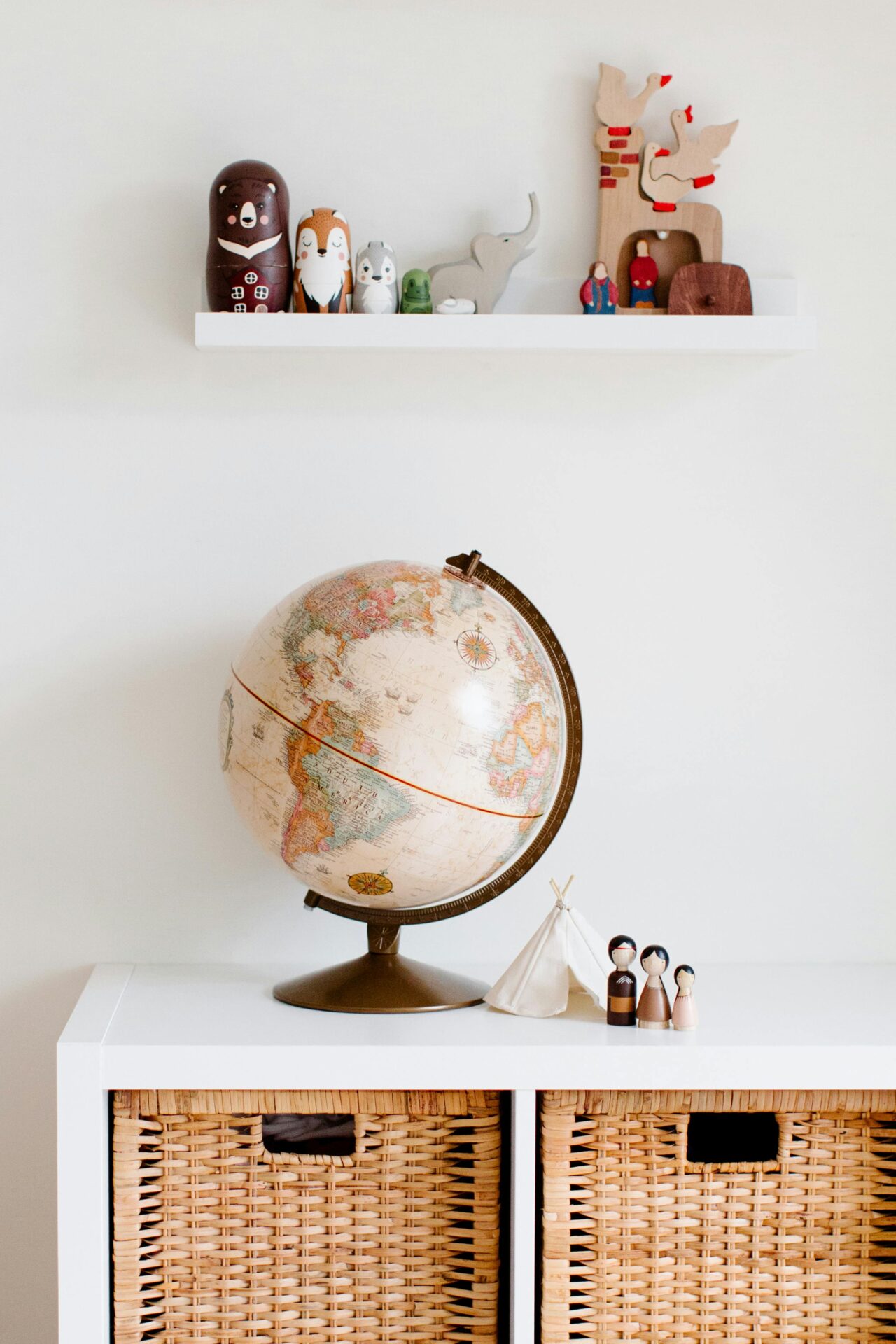 A nursery room setup featuring a globe placed on a white shelf with wicker storage baskets below. A nursery book shelf with various toys and figurines is displayed on a wall-mounted shelf above.