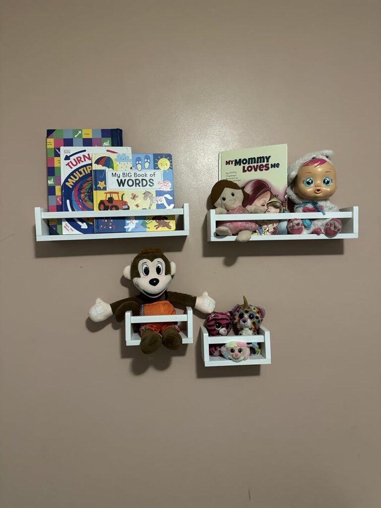 Kids room bookshelf displaying children's books and plush toys, featuring four white shelves arranged in a creative layout on the wall.