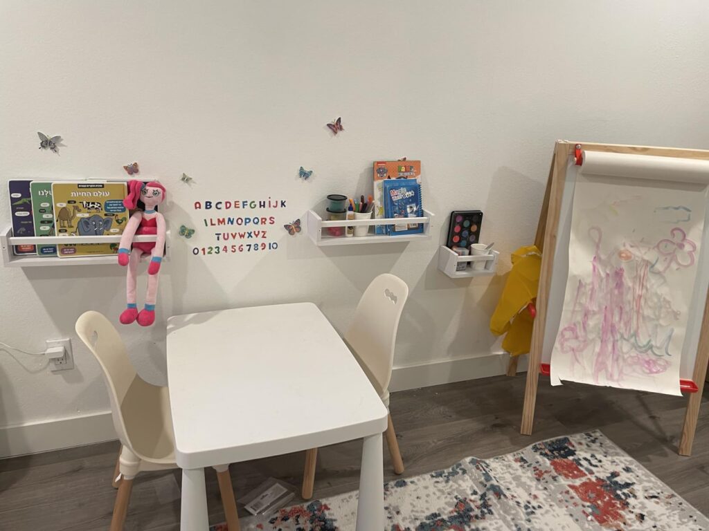 Nursery book Shelves for Wall holding books and toys, mounted above a white children's activity table with a pink doll hanging from one of the shelves.