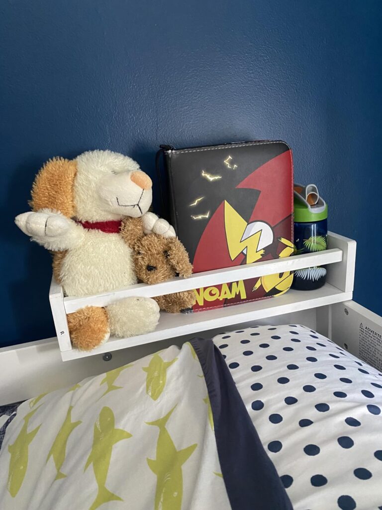 baby book shelf positioned beside the bed, holding stuffed toys, a lunchbox, and a water bottle, making it a convenient storage solution.