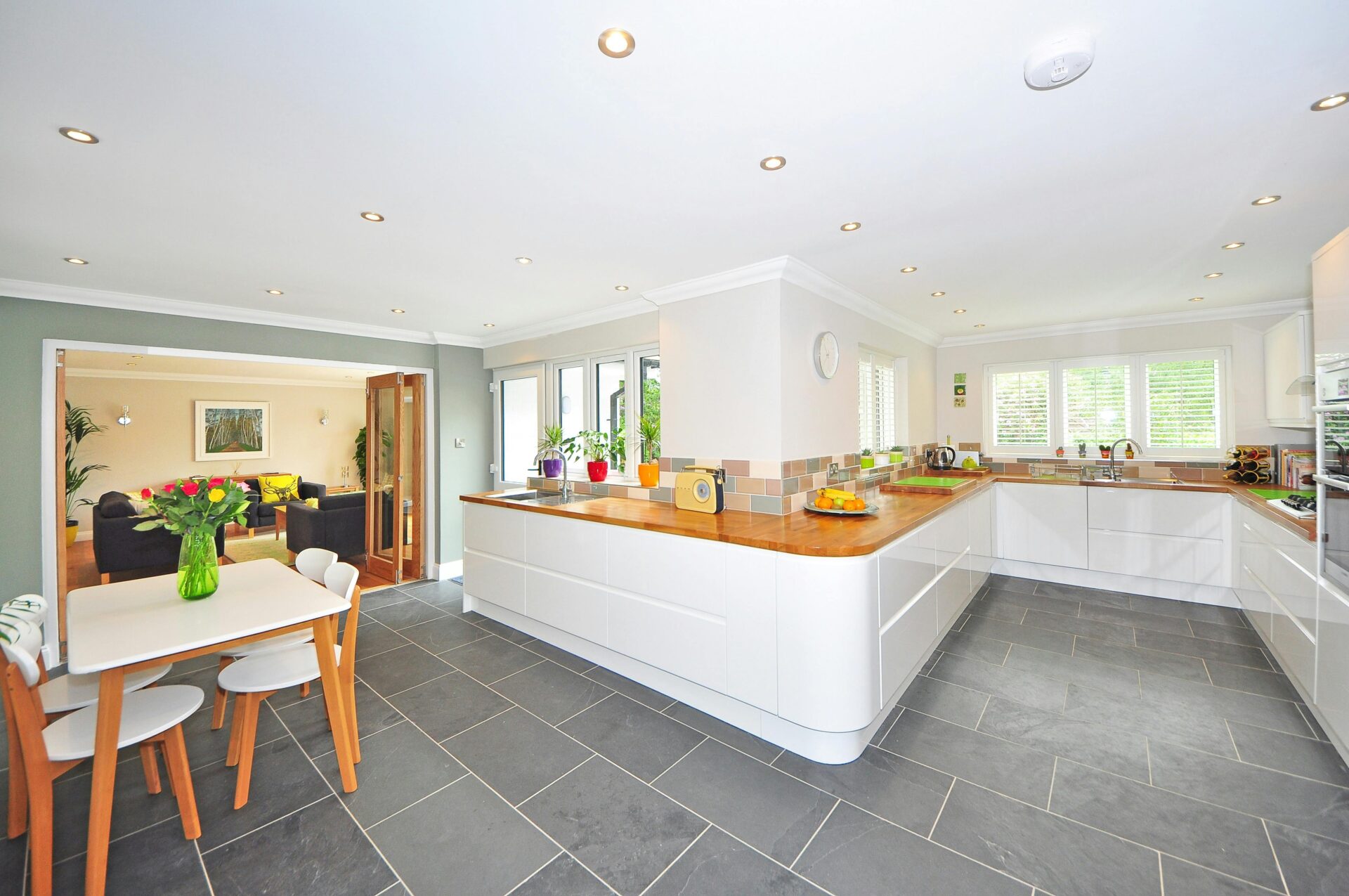 Modern open-concept kitchen and dining area with white cabinets, wooden countertops, and grey tile flooring. The room is well-lit with recessed lighting, and large windows provide natural light.