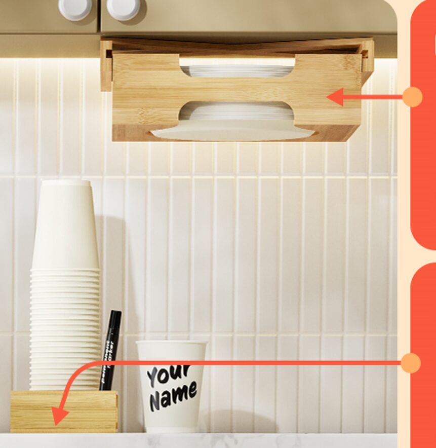 A kitchen setup featuring an under-cabinet bamboo paper plate dispenser holding a stack of plates, with a cut-out for easy access. Below it, a counter displays a stack of white disposable cups next to a marker in a small wooden holder, alongside a cup labeled "Your Name." The background features a tiled backsplash and soft under-cabinet lighting.