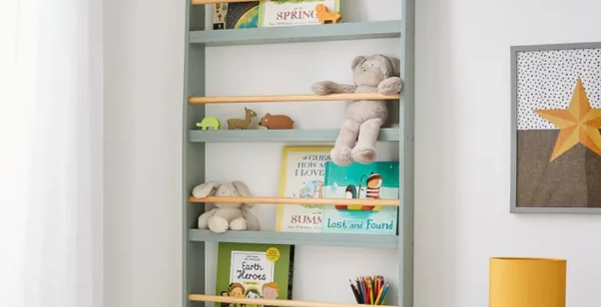 A child's room with a light blue and yellow wooden floating nursery book shelves mounted on the wall, holding books, a stuffed animal, and small toys. A desk with a lamp and colorful pencils is next to the bookshelf, and a star-themed wall art piece is visible.