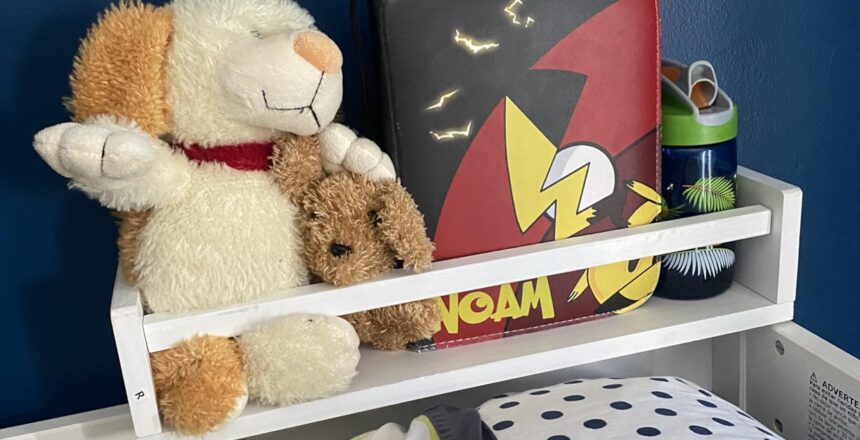 baby book shelf positioned beside the bed, holding stuffed toys, a lunchbox, and a water bottle, making it a convenient storage solution.