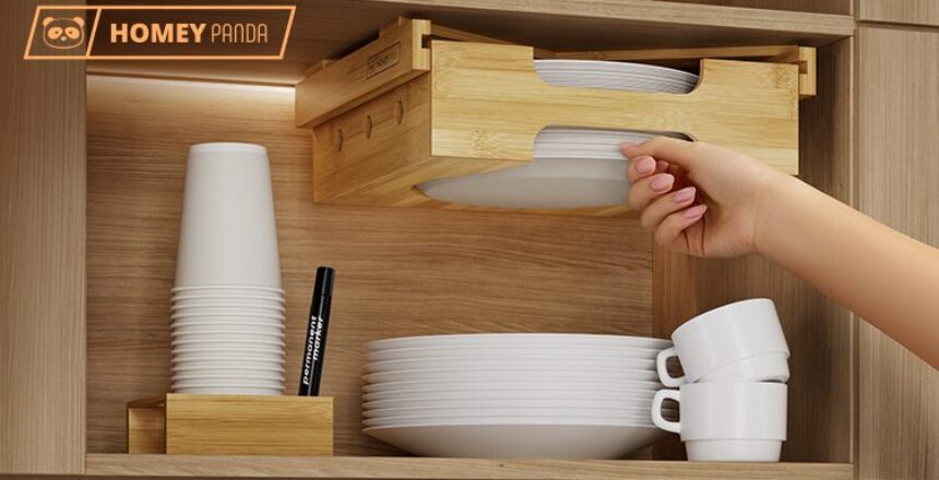 Hand reaching for a plate from a bamboo dispenser mounted in a kitchen cabinet. Next to it, stacked paper cups and a marker are placed in a matching bamboo holder. The setup is branded with 'Homey Panda.'
