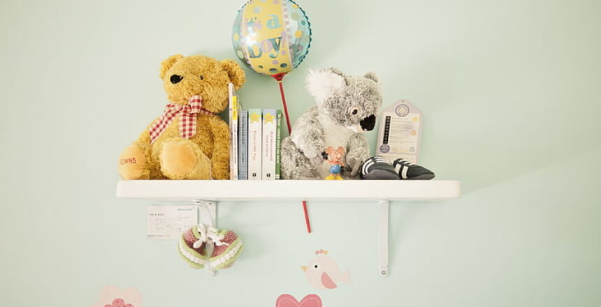 A nursery book shelf mounted on a pastel-colored wall, holding stuffed animals, baby books, and a balloon with 'It's a Boy!' text, creating a cute and cozy atmosphere.